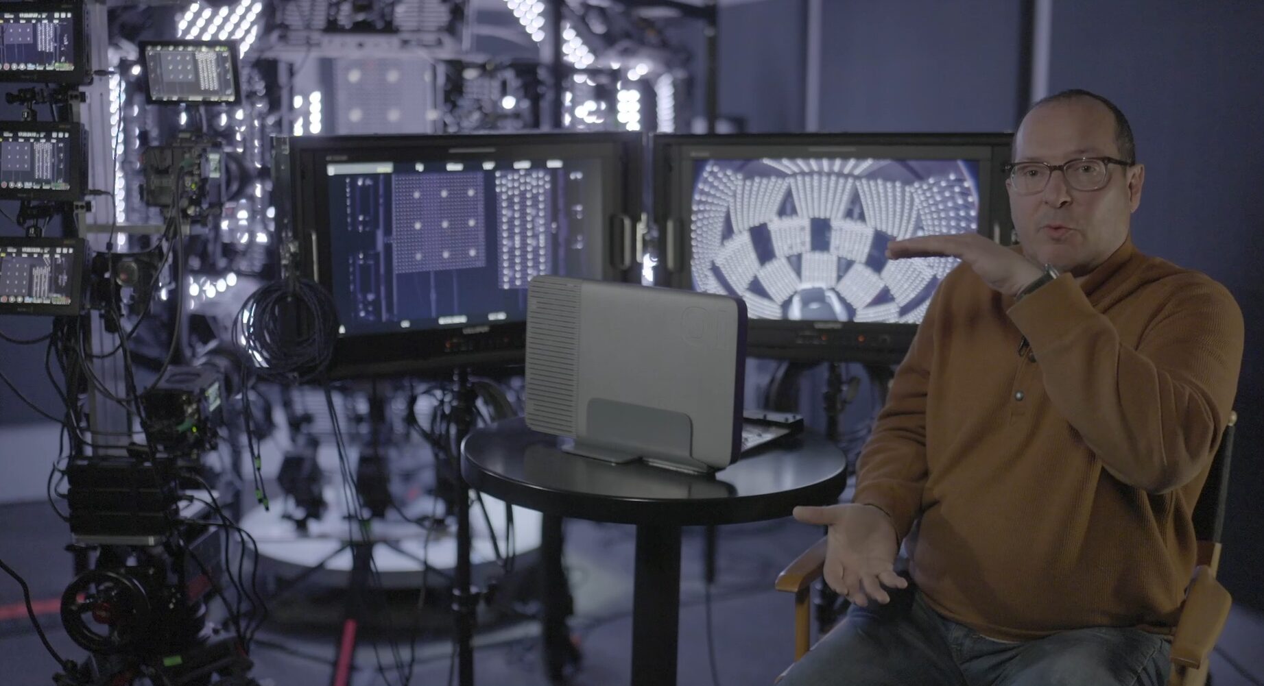 Man sitting in front of monitors and lighting equipment in a visual effects production space and speaking at the camera.